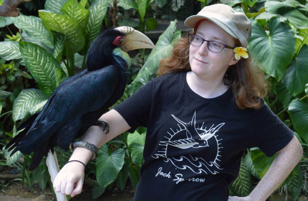 The writer, Avery, holds a large, tropical bird on her arm. She's wearing a brown cap and has a yellow flower tucked behind her ear.